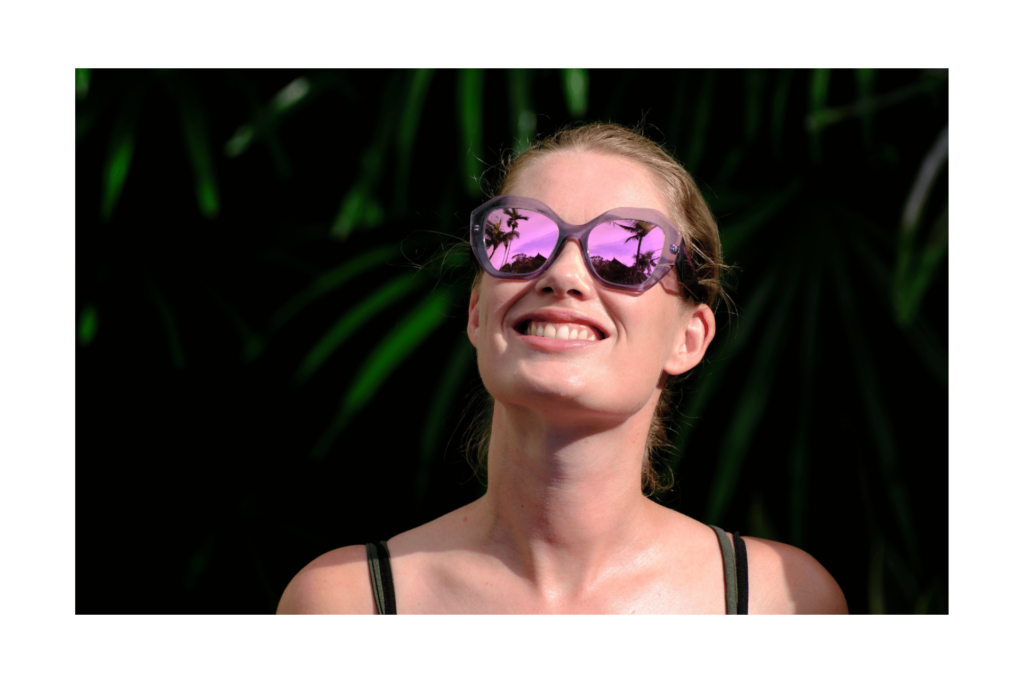 A woman smiles wearing Wassup Beaches sunglasses by Black Shades, a pink frame with pink lenses.