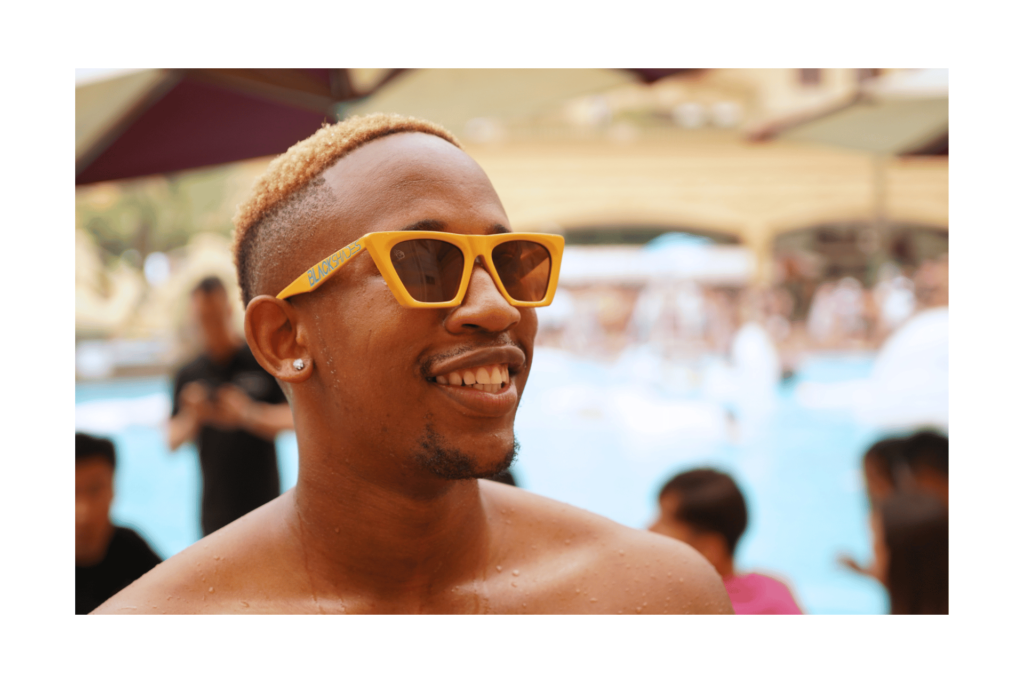 A man smiles wearing Honey Drop sunglasses by Black Shades, a golden frame with amber lenses.