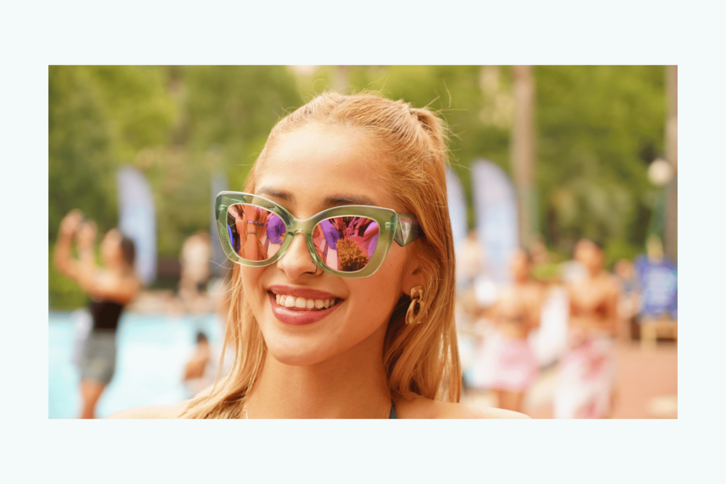 A woman smiles wearing Tempting Teal sunglasses by Black Shades, a teal frame with pink lenses.